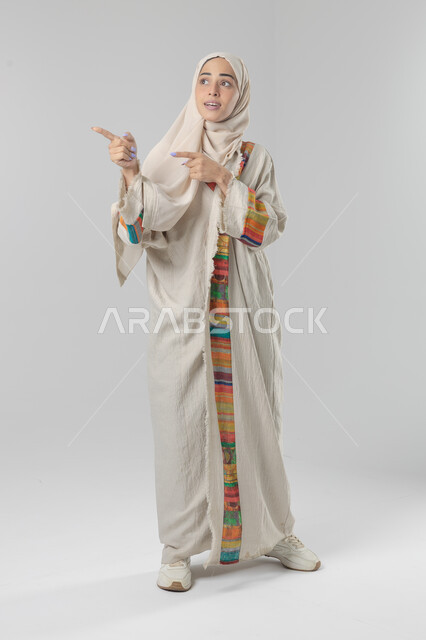 الإشارة إلى عنصر ما, التوضيح والدلالة لمكان محدد, بورتريه لامرأة عربية خليجية سعودية محجبة ترتدي العباءة تشير بيديها على الجانب، تحديد موقع شيء معين, صورة الجسم بالطول الكامل، خلفية بيضاء