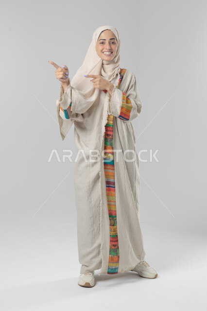 الإشارة إلى عنصر ما, التوضيح والدلالة لمكان محدد, بورتريه لامرأة عربية خليجية سعودية محجبة ترتدي العباءة تشير بيديها على الجانب، تحديد موقع شيء معين, صورة الجسم بالطول الكامل، خلفية بيضاء