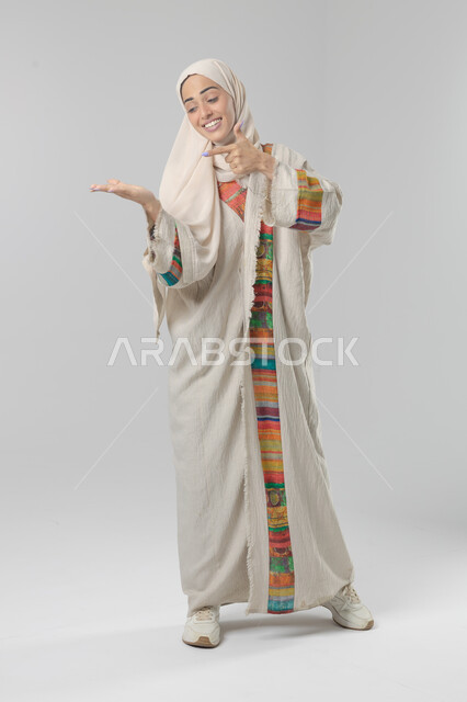 الإشارة إلى عنصر ما, التوضيح والدلالة لمكان محدد, بورتريه لامرأة عربية خليجية سعودية محجبة ترتدي العباءة تشير بيديها على الجانب، تحديد موقع شيء معين, صورة الجسم بالطول الكامل، خلفية بيضاء