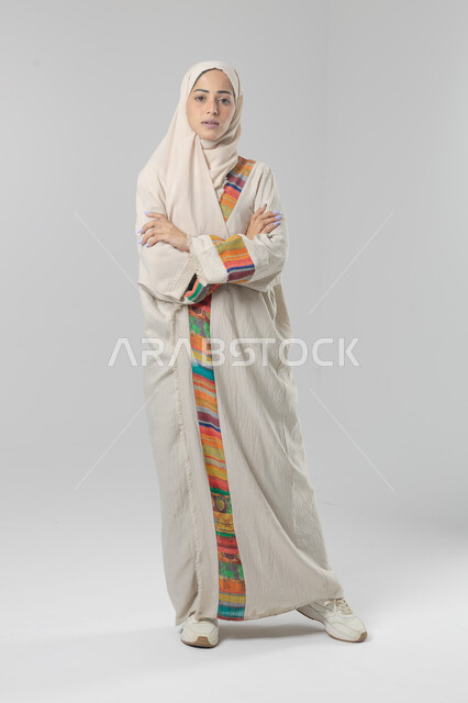 النظر إلى الكاميرا بإيماءات السرور والثقة بالنفس، مفهوم الاناقه و الاهتمام بالمظهر الخارجي، بورتريه لإمرأة عربية خليجية سعودية ترتدي الحجاب والعباءة التقليدية تقف بأيدي متقاطعه، صورة الجسم بالطول الكامل، خلفية بيضاء