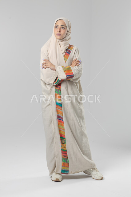 النظر إلى الكاميرا بإيماءات السرور والثقة بالنفس، مفهوم الاناقه و الاهتمام بالمظهر الخارجي، بورتريه لإمرأة عربية خليجية سعودية ترتدي الحجاب والعباءة التقليدية تقف بأيدي متقاطعه، صورة الجسم بالطول الكامل، خلفية بيضاء