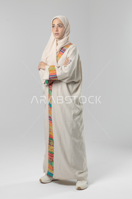Looking at the camera with gestures of pleasure and self-confidence, the concept of elegance and interest in external appearance, a portrait of a Saudi Arabian Gulf woman wearing the hijab and traditional abaya standing with crossed hands, full-length body image, white background