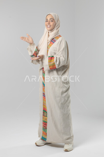 الإشارة إلى عنصر ما, التوضيح والدلالة لمكان محدد, بورتريه لامرأة عربية خليجية سعودية محجبة ترتدي العباءة تشير بيديها على الجانب، تحديد موقع شيء معين, صورة الجسم بالطول الكامل، خلفية بيضاء