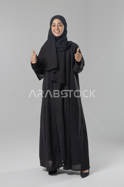 Expressions of approval and acceptance, looking at the camera with gestures of pleasure, satisfaction and admiration, portrait of a smiling veiled Saudi Arabian Gulf woman wearing an abaya, pointing thumbs up in a sign of excellence, full body length, white background
