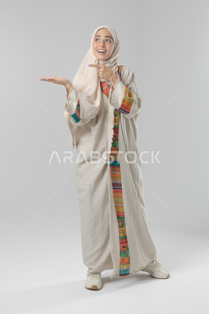 حمل عنصر ما والاشارة اليه, التوضيح والدلالة لمكان محدد, بورتريه لامرأة عربية خليجية سعودية محجبة ترتدي العباءة تشير بيديها على الجانب، تحديد موقع شيء معين, صورة الجسم بالطول الكامل، خلفية بيضاء