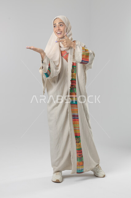 Expressions of approval and acceptance, looking at the camera with gestures of pleasure, satisfaction and admiration, portrait of a smiling veiled Saudi Arabian Gulf woman wearing an abaya, pointing thumbs up in a sign of excellence, full body length, white background