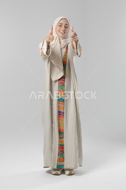 Expressions of approval and acceptance, looking at the camera with gestures of pleasure, satisfaction and admiration, portrait of a smiling veiled Saudi Arabian Gulf woman wearing an abaya, pointing thumbs up in a sign of excellence, full body length, white background