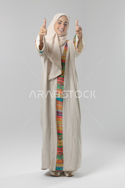 تعابير الموافقة والقبول, النظر الى الكاميرا بإيماءات السرور الرضى والاعجاب، بورتريه لامرأة عربيه خليجية سعودية مبتسمة محجبة ترتدي العباءه تشير باصابع الابهام الى الاعلى بعلامه الامتياز، الطول الكامل للجسم، خلفية بيضاء