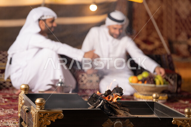 كشته بر وتخييم شبابيه في صحراء الامارات, ديكور وزينة شهر رمضان, تقديم الفاكهة الطازجة للضيف، حوارات ونقاشات بين الاصدقاء, رجلان عربيان خليجيان اماراتيان يرتديان الكندوره والغتره يجلسان في خيمه شعبيه, حسن الاستقبال وكرم الضيافه، أجواء ليلية أمام شعلة الحطب