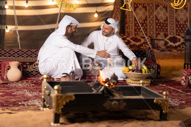 كشته بر وتخييم شبابيه في صحراء الامارات, ديكور وزينة شهر رمضان, تقديم الفاكهة الطازجة للضيف، حوارات ونقاشات بين الاصدقاء, رجلان عربيان خليجيان اماراتيان يرتديان الكندوره والغتره يجلسان في خيمه شعبيه, حسن الاستقبال وكرم الضيافه، أجواء ليلية أمام شعلة الحطب
