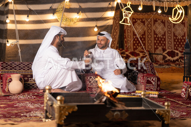 تقديم القهوة العربية الساخنة للضيف، حوارات ونقاشات بين الاصدقاء, رجلان عربيان خليجيان اماراتيان يرتديان الكندوره والغتره يجلسان في خيمه شعبيه, حسن الاستقبال وكرم الضيافه، ديكور وزينة شهر رمضان, كشته بر وتخييم شبابيه في صحراء الامارات, أجواء ليلية أمام شعلة الحطب