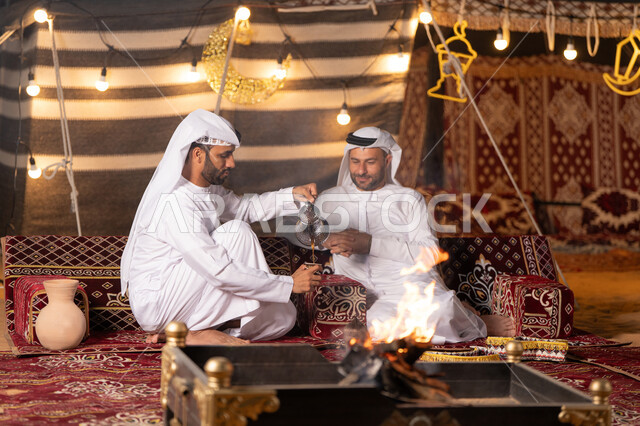ديكور وزينة شهر رمضان, تقديم القهوة العربية الساخنة للضيف، حوارات ونقاشات بين الاصدقاء, رجلان عربيان خليجيان اماراتيان يرتديان الكندوره والغتره يجلسان في خيمه شعبيه, حسن الاستقبال وكرم الضيافه، كشته بر وتخييم شبابيه في صحراء الامارات, أجواء ليلية أمام شعلة الحطب