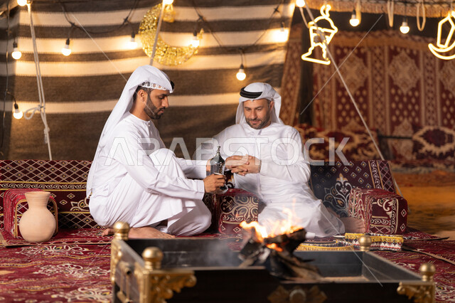تقديم القهوة العربية الساخنة للضيف، حوارات ونقاشات بين الاصدقاء, رجلان عربيان خليجيان اماراتيان يرتديان الكندوره والغتره يجلسان في خيمه شعبيه, حسن الاستقبال وكرم الضيافه، ديكور وزينة شهر رمضان, كشته بر وتخييم شبابيه في صحراء الامارات, أجواء ليلية أمام شعلة الحطب