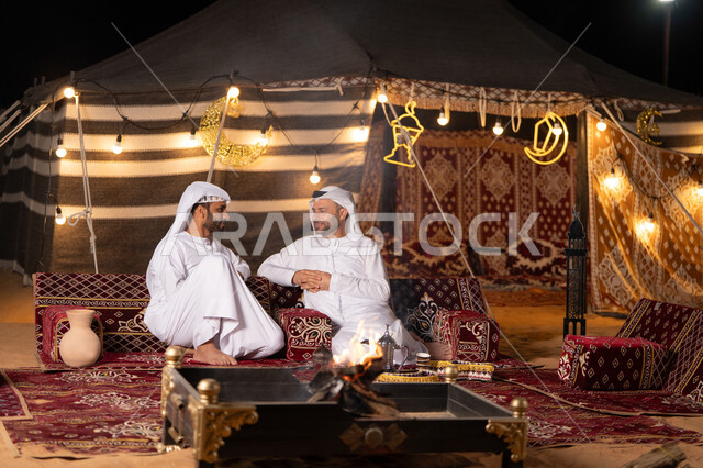 Dialogues and discussions between friends, two Emirati Gulf Arab men wearing kandura and ghutra sitting in a traditional Arab tent, warm welcome and generous hospitality, Ramadan decorations and lighting, a youth camp in the UAE desert, a quiet night atmosphere in front of a firewood flame