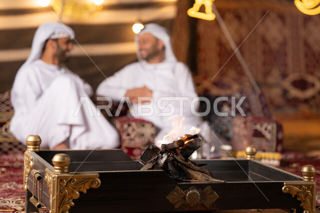 زينة وديكور وإضاءة شهر رمضان, حوار ونقاش بين الاصدقاء, رجلان عربيان خليجيان اماراتيان يرتديان الكندوره والغتره يجلسان في خيمه عربيه شعبيه, حسن الاستقبال وكرم الضيافه، كشته بر وتخييم شبابيه في صحراء الامارات, أجواء ليلية هادئة، صورة مقربة لموقد الحطب المشتعل