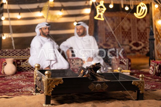 حوارات ونقاشات بين الاصدقاء, رجلان عربيان خليجيان اماراتيان يرتديان الكندوره والغتره يجلسان في خيمه عربيه شعبيه, حسن الاستقبال وكرم الضيافه، ديكور وزينة وإضاءة شهر رمضان, كشته بر وتخييم شبابيه في صحراء الامارات, أجواء ليلية هادئة أمام شعلة الحطب