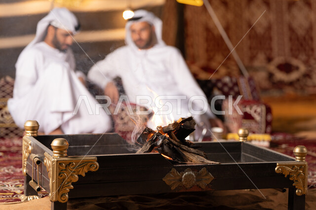 زينة وديكور وإضاءة شهر رمضان, حوار ونقاش بين الاصدقاء, رجلان عربيان خليجيان اماراتيان يرتديان الكندوره والغتره يجلسان في خيمه عربيه شعبيه, حسن الاستقبال وكرم الضيافه، كشته بر وتخييم شبابيه في صحراء الامارات, أجواء ليلية هادئة، صورة مقربة لموقد الحطب المشتعل