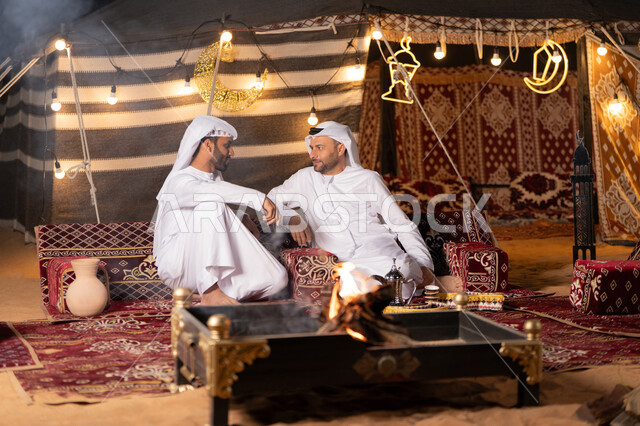 Dialogues and discussions between friends, two Emirati Gulf Arab men wearing kandura and ghutra sitting in a traditional Arab tent, warm welcome and generous hospitality, Ramadan decorations and lighting, a youth camp in the UAE desert, a quiet night atmosphere in front of a firewood flame