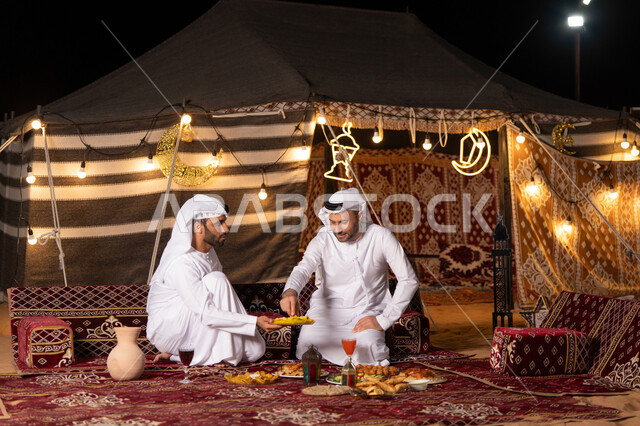 مائدة مليئة بأصناف الطعام والحلوى الرمضانية، زينة وإضاءة شهر رمضان, رجلان عربيان خليجيان اماراتيان يرتديان الكندوره والغتره يجلسان في خيمه عربيه شعبيه, حسن الاستقبال وكرم الضيافه، أجواء ليلية هادئة في كشتة بر وتخييم شبابيه في صحراء الامارات