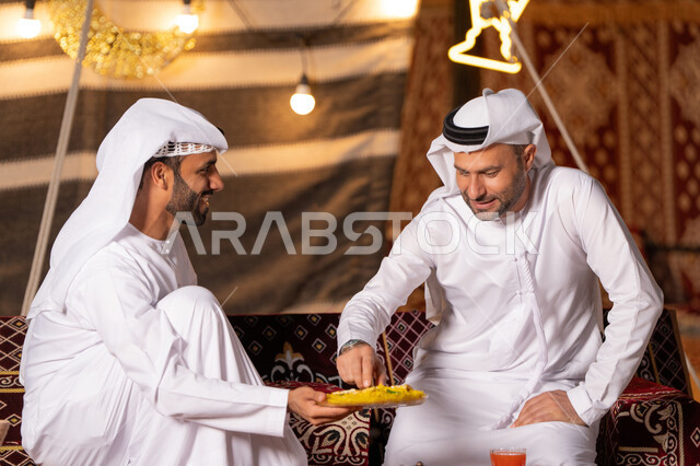 كشتة بر وتخييم شبابيه في صحراء الامارات, زينة وإضاءة شهر رمضان, تقديم الحلوى الرمضانية للضيف، رجلان عربيان خليجيان اماراتيان يرتديان الكندوره والغتره يجلسان في خيمه عربيه شعبيه, حسن الاستقبال وكرم الضيافه