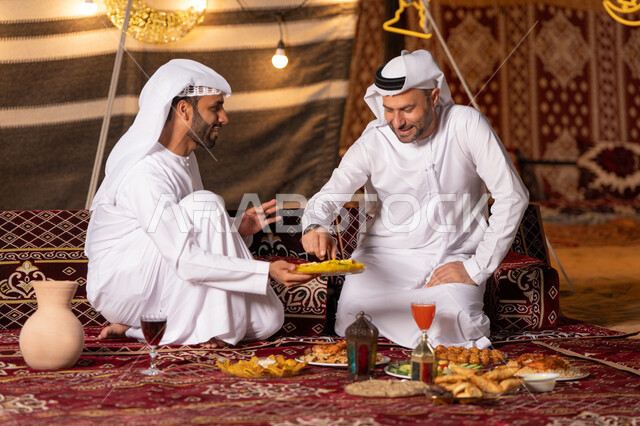 زينة وإضاءة شهر رمضان, مائدة مليئة بأصناف الطعام والحلوى الرمضانية، رجلان عربيان خليجيان اماراتيان يرتديان الكندوره والغتره يجلسان في خيمه عربيه شعبيه, حسن الاستقبال وكرم الضيافه، أجواء ليلية هادئة في كشتة بر وتخييم شبابيه في صحراء الامارات