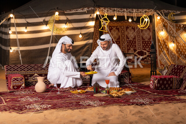 مائدة مليئة بأصناف الطعام والحلوى الرمضانية، زينة وإضاءة شهر رمضان, رجلان عربيان خليجيان اماراتيان يرتديان الكندوره والغتره يجلسان في خيمه عربيه شعبيه, حسن الاستقبال وكرم الضيافه، أجواء ليلية هادئة في كشتة بر وتخييم شبابيه في صحراء الامارات