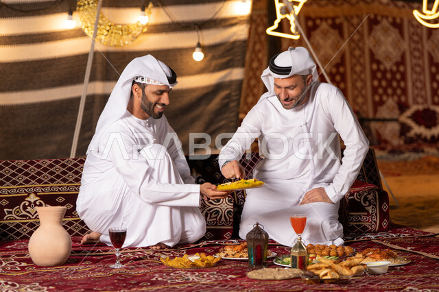 زينة وإضاءة شهر رمضان, مائدة مليئة بأصناف الطعام والحلوى الرمضانية، رجلان عربيان خليجيان اماراتيان يرتديان الكندوره والغتره يجلسان في خيمه عربيه شعبيه, حسن الاستقبال وكرم الضيافه، أجواء ليلية هادئة في كشتة بر وتخييم شبابيه في صحراء الامارات