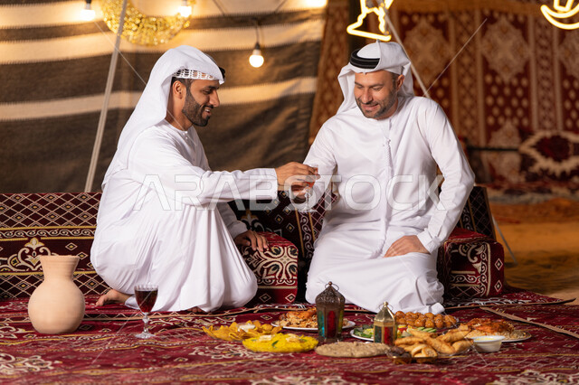 مائدة مليئة بأصناف الطعام والحلوى الرمضانية، زينة وإضاءة شهر رمضان, رجلان عربيان خليجيان اماراتيان يرتديان الكندوره والغتره يجلسان في خيمه عربيه شعبيه, حسن الاستقبال وكرم الضيافه، أجواء ليلية هادئة في كشتة بر وتخييم شبابيه في صحراء الامارات