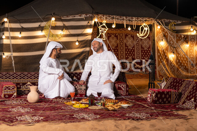 زينة وإضاءة شهر رمضان, مائدة مليئة بأصناف الطعام والحلوى الرمضانية، رجلان عربيان خليجيان اماراتيان يرتديان الكندوره والغتره يجلسان في خيمه عربيه شعبيه, حسن الاستقبال وكرم الضيافه، أجواء ليلية هادئة في كشتة بر وتخييم شبابيه في صحراء الامارات