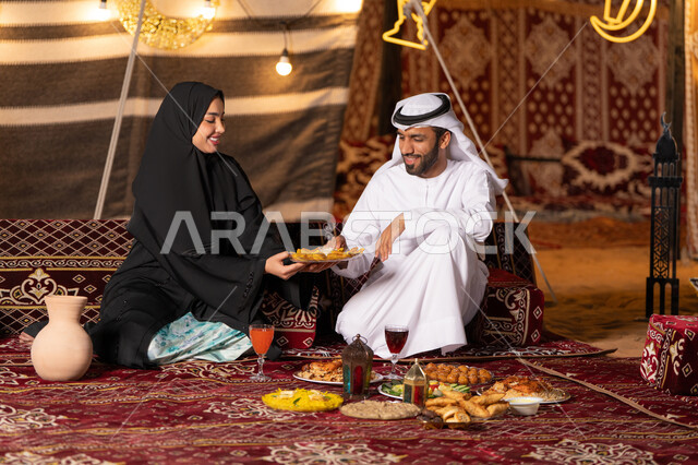 A table full of Ramadan food and sweets, decorations and lighting for the holy month of Ramadan, a smiling Emirati Gulf Arab couple sitting in a traditional Arab tent with happy gestures, a quiet night atmosphere in a desert outing and family camping in the UAE desert, spending a fun time with the family
