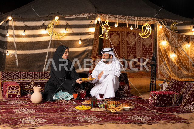 مائدة مليئة بأصناف الطعام والحلوى الرمضانية، زينة وإضاءة شهر رمضان المبارك, زوجان عربيان خليجيان اماراتيان مبتسمان يجلسان في خيمه عربيه شعبيه بإيماءات السعادة, أجواء ليلية هادئة في كشتة بر وتخييم عائلية في صحراء الامارات، قضاء وقت ممتع مع الأسرة