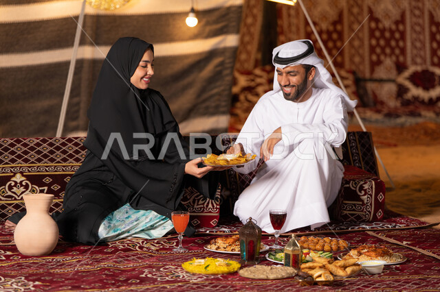 أجواء ليلية هادئة في كشتة بر وتخييم عائلية في صحراء الامارات، قضاء وقت ممتع مع الأسرة، مائدة مليئة بأصناف الطعام والحلوى الرمضانية، زينة وإضاءة شهر رمضان المبارك, زوجان عربيان خليجيان اماراتيان مبتسمان يجلسان في خيمه عربيه شعبيه بإيماءات السعادة