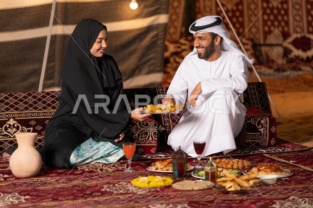 A quiet night atmosphere in a family camping trip in the UAE desert, spending a pleasant time with the family, a table full of Ramadan food and sweets, decorations and lighting for the holy month of Ramadan, a smiling Emirati Gulf Arab couple sitting in a traditional Arab tent with gestures of happiness