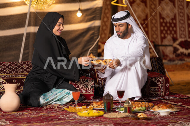 قضاء وقت ممتع مع الأسرة، مائدة مليئة بأصناف الطعام والحلوى الرمضانية، زينة وإضاءة شهر رمضان المبارك, زوجان عربيان خليجيان اماراتيان مبتسمان يجلسان في خيمه عربيه شعبيه بإيماءات السعادة, أجواء ليلية هادئة في كشتة بر وتخييم عائلية في صحراء الامارات