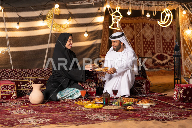 مائدة مليئة بأصناف الطعام والحلوى الرمضانية، زينة وإضاءة شهر رمضان المبارك, زوجان عربيان خليجيان اماراتيان مبتسمان يجلسان في خيمه عربيه شعبيه بإيماءات السعادة, أجواء ليلية هادئة في كشتة بر وتخييم عائلية في صحراء الامارات، قضاء وقت ممتع مع الأسرة