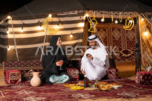 مائدة مليئة بأصناف الطعام والحلوى الرمضانية، زينة وإضاءة شهر رمضان المبارك, زوجان عربيان خليجيان اماراتيان مبتسمان يجلسان في خيمه عربيه شعبيه بإيماءات السعادة, أجواء ليلية هادئة في كشتة بر وتخييم عائلية في صحراء الامارات، قضاء وقت ممتع مع الأسرة