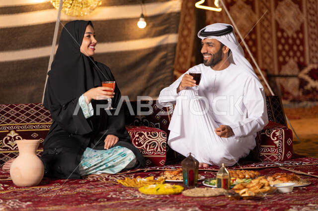 أجواء ليلية هادئة في كشتة بر وتخييم عائلية في صحراء الامارات، قضاء وقت ممتع مع الأسرة، مائدة مليئة بأصناف الطعام والحلوى الرمضانية، زينة وإضاءة شهر رمضان المبارك, زوجان عربيان خليجيان اماراتيان مبتسمان يجلسان في خيمه عربيه شعبيه بإيماءات السعادة