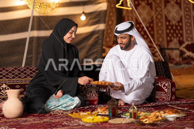 قضاء وقت ممتع مع الأسرة، مائدة مليئة بأصناف الطعام والحلوى الرمضانية، زينة وإضاءة شهر رمضان المبارك, زوجان عربيان خليجيان اماراتيان مبتسمان يجلسان في خيمه عربيه شعبيه بإيماءات السعادة, أجواء ليلية هادئة في كشتة بر وتخييم عائلية في صحراء الامارات