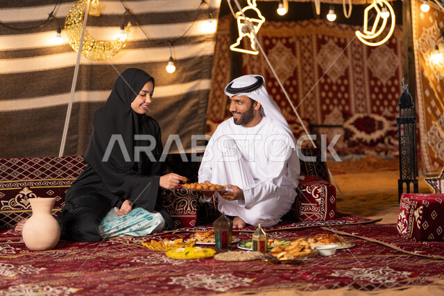 إفطار رمضاني عائلي، أجواء وطقوس دينية إسلامية، مائدة إفطار مليئة بالمأكولات اللذيذة والحلويات، زوجان عربيان خليجيان اماراتيان يرتديان الزي التقليدي يتشاركان الحلوي الرمضانية الشرقية بايماءات السعادة و السرور، التخييم في الصحراء، زينة وإضاءة الأعياد والمناسبات