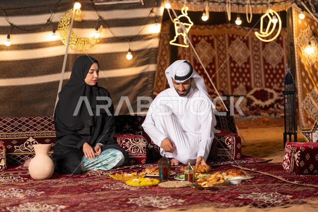 مائدة إفطار مليئة بالمأكولات اللذيذة والحلويات، إفطار رمضاني عائلي، أجواء وطقوس دينية إسلامية، زوجان عربيان خليجيان اماراتيان يرتديان الزي التقليدي يقومان بتناول الافطار بايماءات السعادة و السرور، التخييم في الصحراء، زينة وإضاءة الأعياد والمناسبات