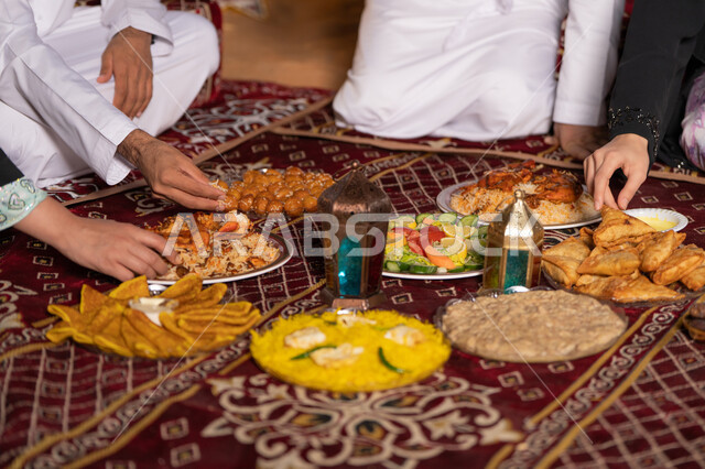 زينة وأضواء وفوانيس الاحتفال بقدوم شهر الخير، المأكولات العربية الشرقية, سفرة مليئة بالأصناف والمقبلات الشهية, صورة مقربة ليد عائله عربيه خليجيه اماراتية يقومون بتناول الافطار وقت اذان المغرب ، إفطار رمضاني عائلي
