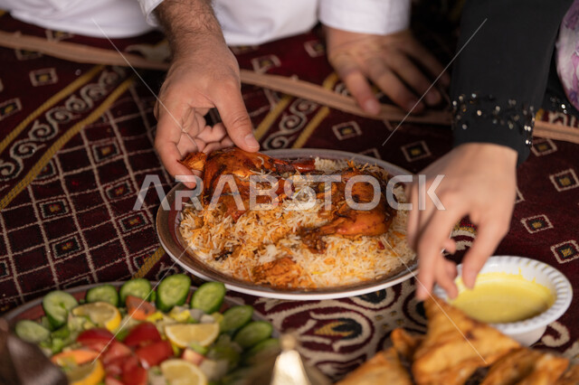حلول موعد الإفطار عند اذان المغرب، المأكولات العربية الشرقية, زينة وأضواء وفوانيس الاحتفال بقدوم شهر الخير, سفرة مليئة بالأصناف والمقبلات الشهية, صورة مقربة ليد زوجان عربيان خليجان اماراتيان يقومان بتناول الافطار الشهي