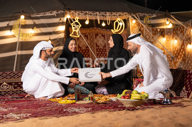 مفهوم الترابط الأسري, الاستمتاع بتناول الإفطار بالهواء الطلق مع الأهل, عائلة عربية خليجية اماراتيه يرتدون الثوب التقليدي يتناولون الافطار, رجل عربي يقوم بتقديم صندوق التمر الرمضاني , ايماءات السعاده و السرور , زينه و ديكور شهر رمضان, التخييم في الصحراء