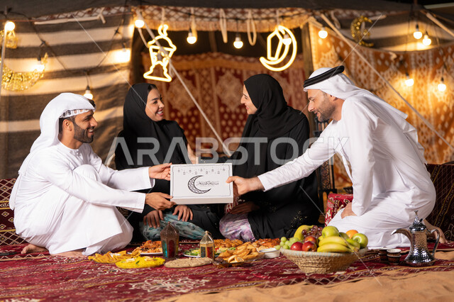 الاستمتاع بتناول الإفطار بالهواء الطلق مع الأهل, مفهوم الترابط الأسري, عائلة عربية خليجية اماراتيه يرتدون الثوب التقليدي يتناولون الافطار, رجل عربي يقوم بتقديم صندوق التمر الرمضاني , ايماءات السعاده و السرور , زينه و ديكور شهر رمضان, التخييم في الصحراء