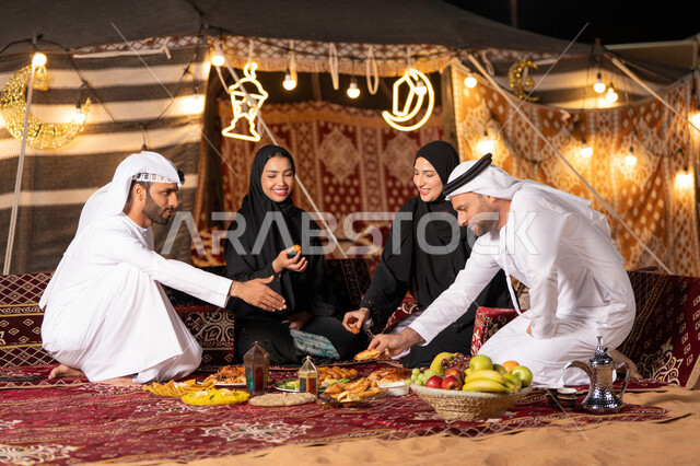مائدة إفطار مليئة بأصناف المأكولات الشهية، التخييم في الصحراء وقت الافطار، مفهوم الضيافة العربية وحسن استقبال الضيوف، عائلة عربية خليجية اماراتيه يرتدون الثوب التقليدي يجلسون على مائدة الطعام, أجواء شهر رمضان المبارك , أصناف متنوعة من المأكولات