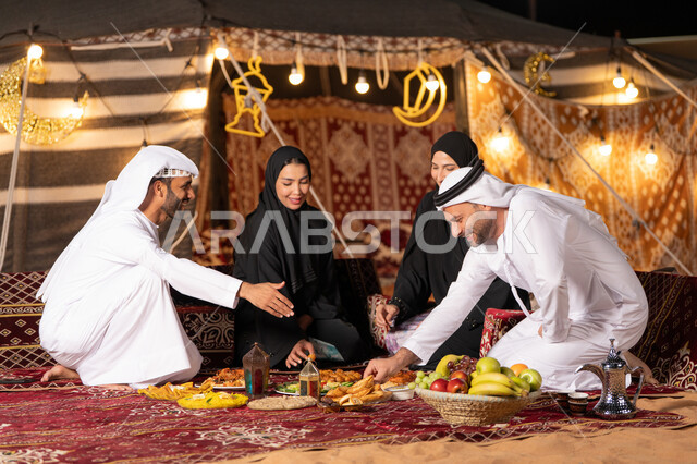 مائدة إفطار مليئة بأصناف المأكولات الشهية، التخييم في الصحراء وقت الافطار، مفهوم الضيافة العربية وحسن استقبال الضيوف، عائلة عربية خليجية اماراتيه يرتدون الثوب التقليدي يجلسون على مائدة الطعام, أجواء شهر رمضان المبارك , أصناف متنوعة من المأكولات
