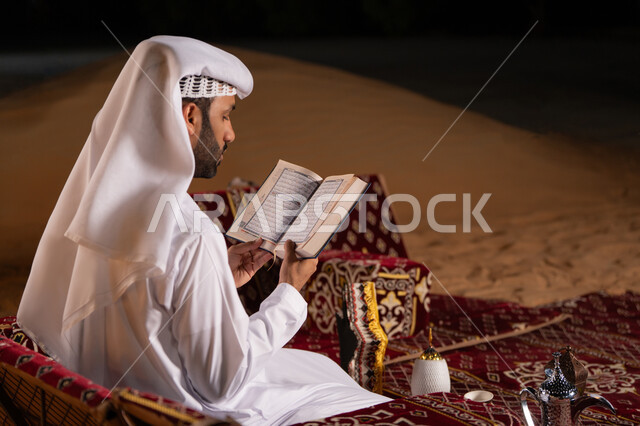 Reciting and memorizing the Holy Quran in the spiritual atmosphere of Ramadan, a side view of an Emirati Gulf Arab man wearing a white kandura and ghutra reading the Holy Quran, camping in the desert at night, getting closer to God in the holy month of Ramadan, striving in worship and performing religious duties.