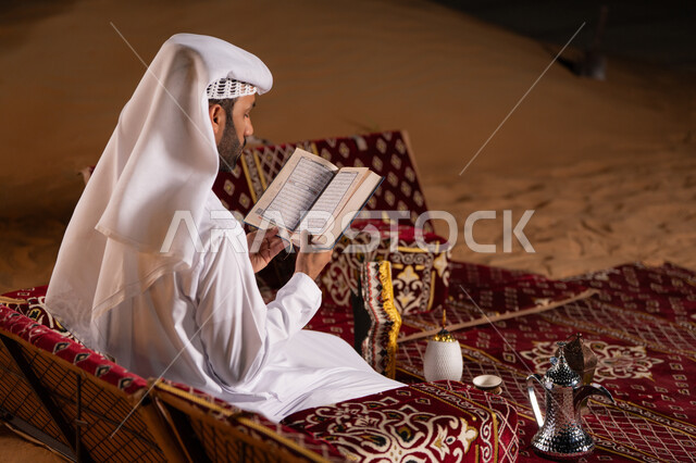 Reciting and memorizing the Holy Quran in the spiritual atmosphere of Ramadan, a side view of an Emirati Gulf Arab man wearing a white kandura and ghutra reading the Holy Quran, camping in the desert at night, getting closer to God in the holy month of Ramadan, striving in worship and performing religious duties.