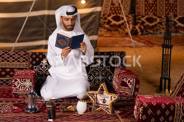 التعبد بتلاوة القران وحفظ وتفسير كلامه، رجل عربي خليجي اماراتي يرتدي الكندوره والغتره البيضاء يحمل المصحف الشريف، الجو الديني الروحاني, العبادة والتقرب إلى رب العالمين، كشته بر شبابيه، التخييم في اجواء رمضانيه، خيمه عربيه مليىْه بديكورات شهر رمضان