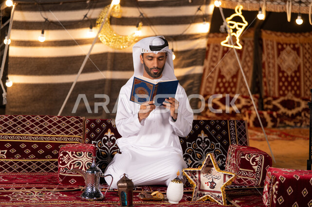 التعبد بتلاوة القران وحفظ وتفسير كلامه، رجل عربي خليجي اماراتي يرتدي الكندوره والغتره البيضاء يحمل المصحف الشريف، الجو الديني الروحاني, العبادة والتقرب إلى رب العالمين، كشته بر شبابيه، التخييم في اجواء رمضانيه، خيمه عربيه مليىْه بديكورات شهر رمضان